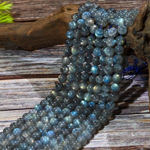 May include: Strands of round, iridescent labradorite beads in shades of gray and blue. The beads are strung together, creating a flowing, textured appearance. The beads are displayed on a wooden surface.