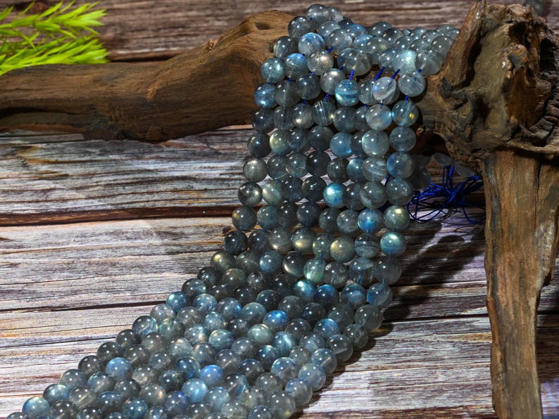 May include: Strands of round, iridescent labradorite beads in shades of gray and blue. The beads are strung together, creating a flowing, textured appearance. The beads are displayed on a wooden surface.