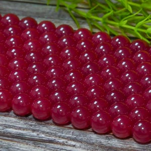 Peut inclure: Gros plan sur une collection de perles rondes, polies, rouge fonc&eacute;. Les perles sont dispos&eacute;es de pr&egrave;s, refl&eacute;tant la lumi&egrave;re et cr&eacute;ant un aspect brillant. Du feuillage vert est visible en arri&egrave;re-plan, ajoutant un contraste naturel.