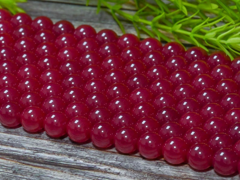Peut inclure: Gros plan sur une collection de perles rondes, polies, rouge fonc&eacute;. Les perles sont dispos&eacute;es de pr&egrave;s, refl&eacute;tant la lumi&egrave;re et cr&eacute;ant un aspect brillant. Du feuillage vert est visible en arri&egrave;re-plan, ajoutant un contraste naturel.