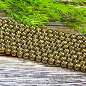 May include: A strand of gold-toned round beads, likely made of hematite, arranged in rows on a wooden surface. The beads are smooth and shiny, reflecting light.