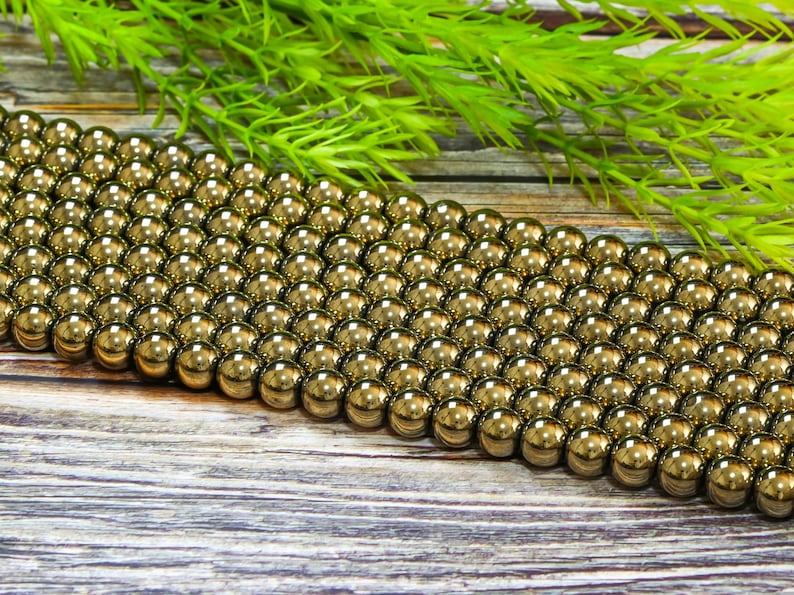 May include: A strand of gold-toned round beads, likely made of hematite, arranged in rows on a wooden surface. The beads are smooth and shiny, reflecting light.