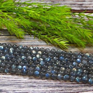 May include: Strands of round, gray labradorite gemstone beads with blue and iridescent flashes. The beads are displayed on a wooden surface with green foliage in the background. These beads are perfect for jewelry making.