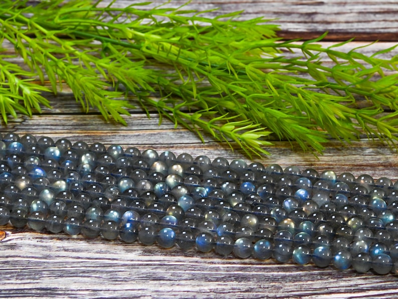 May include: Strands of round, gray labradorite gemstone beads with blue and iridescent flashes. The beads are displayed on a wooden surface with green foliage in the background. These beads are perfect for jewelry making.