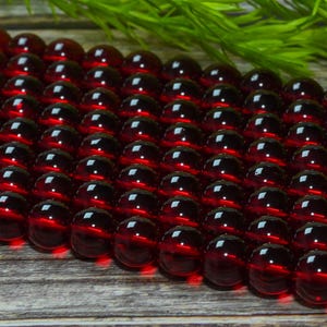 May include: A collection of deep red, spherical beads arranged in multiple parallel rows. Each bead features a smooth, polished, and translucent surface that reflects light, creating a consistent circular highlight on the top of every individual bead.