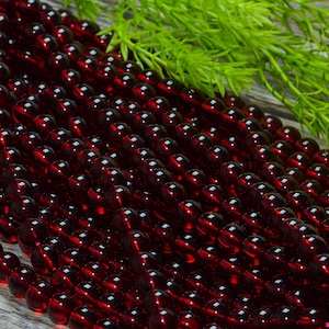 May include: A collection of deep red, translucent, spherical beads arranged in multiple parallel strands. The beads have a smooth, polished surface that reflects light, creating small highlights across the dark crimson, circular forms.