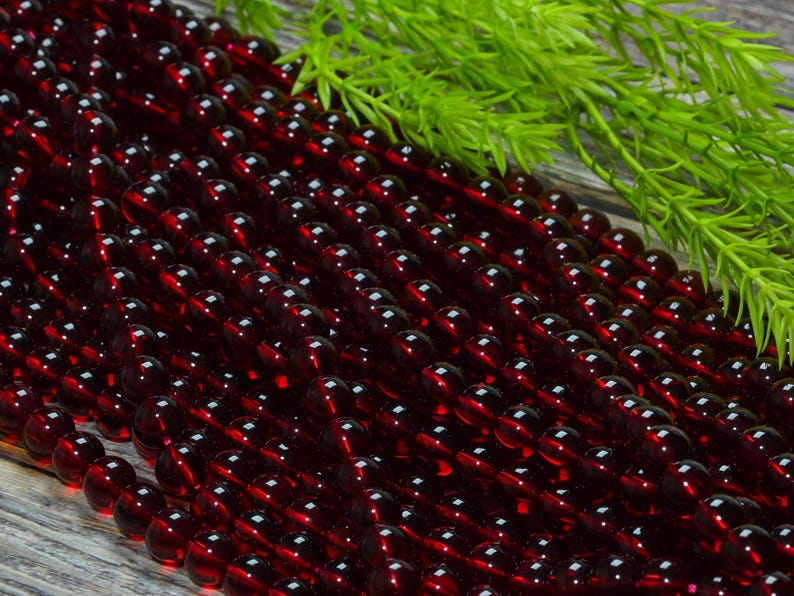 May include: A collection of deep red, translucent, spherical beads arranged in multiple parallel strands. The beads have a smooth, polished surface that reflects light, creating small highlights across the dark crimson, circular forms.