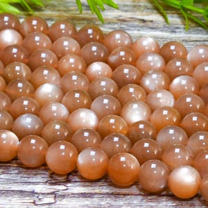 May include: A strand of round, peach-colored sunstone beads. The beads have a pearlescent sheen and are arranged in a close-up view.