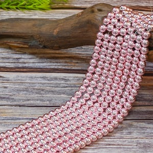 Op de afbeelding: Een streng roze glazen kralen, elke kraal is rond en heeft een glanzende, metallic afwerking. De kralen zijn in een rij op een houten oppervlak gerangschikt.