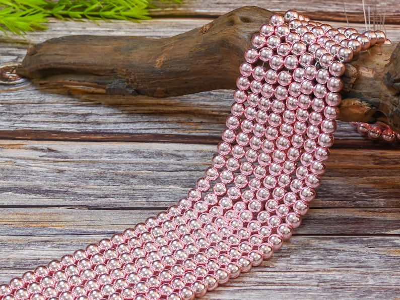 Op de afbeelding: Een streng roze glazen kralen, elke kraal is rond en heeft een glanzende, metallic afwerking. De kralen zijn in een rij op een houten oppervlak gerangschikt.