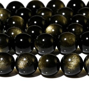 Könnte beinhalten: Eine Kette aus schwarzen Perlen mit goldenen Sprenkeln. Die Perlen sind rund und glatt und auf einer durchsichtigen Schnur aufgefädelt.