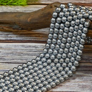 May include: A strand of silver-toned round beads, likely made of hematite, arranged in a cascading pattern on a wooden surface.