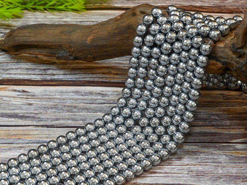 May include: A strand of silver-toned round beads, likely made of hematite, arranged in a cascading pattern on a wooden surface.