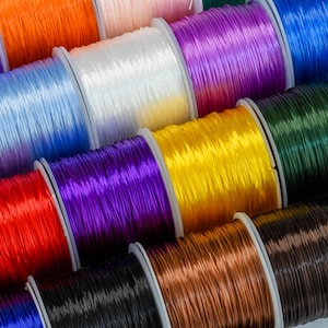 Könnte beinhalten: Nahaufnahme von Spulen mit farbigem Gummiband. Die Bänder sind fest gewickelt und in einer Reihe angeordnet. Die Farben umfassen Pink, Blau, Weiß, Lila, Gelb, Grün, Braun, Schwarz und Rot.