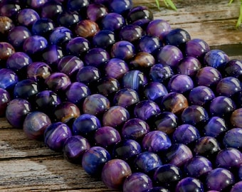 Puede incluir: Una hebra de cuentas de ojo de tigre morado. Las cuentas son redondas y tienen un acabado liso y pulido. Son de color morado oscuro con vetas marrones y doradas.