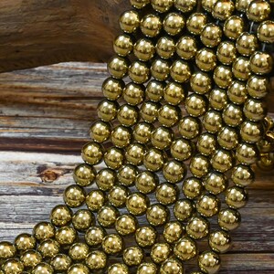 May include: A strand of gold-toned round beads. The beads are smooth and shiny, and they are arranged in a cascading pattern. The beads are on a wooden surface.