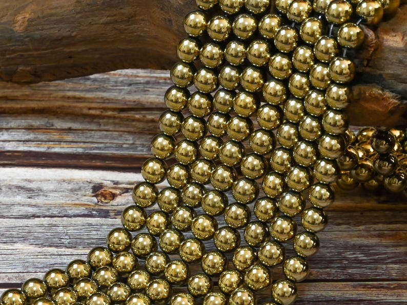 May include: A strand of gold-toned round beads. The beads are smooth and shiny, and they are arranged in a cascading pattern. The beads are on a wooden surface.
