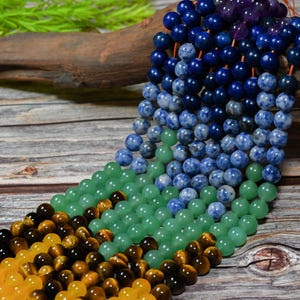 May include: Strands of colorful gemstone beads in shades of yellow, brown, green, blue, and purple. The beads are round and appear to be strung together, with a wooden background.