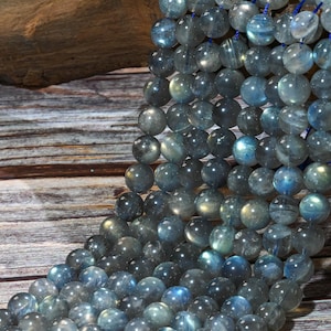 May include: Strands of round, polished labradorite beads in shades of gray, blue, and green. The beads are strung together, showcasing their iridescent quality. The background features a wooden surface.