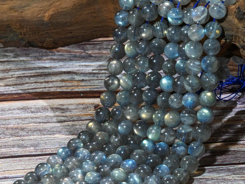 May include: Strands of round, polished labradorite beads in shades of gray, blue, and green. The beads are strung together, showcasing their iridescent quality. The background features a wooden surface.