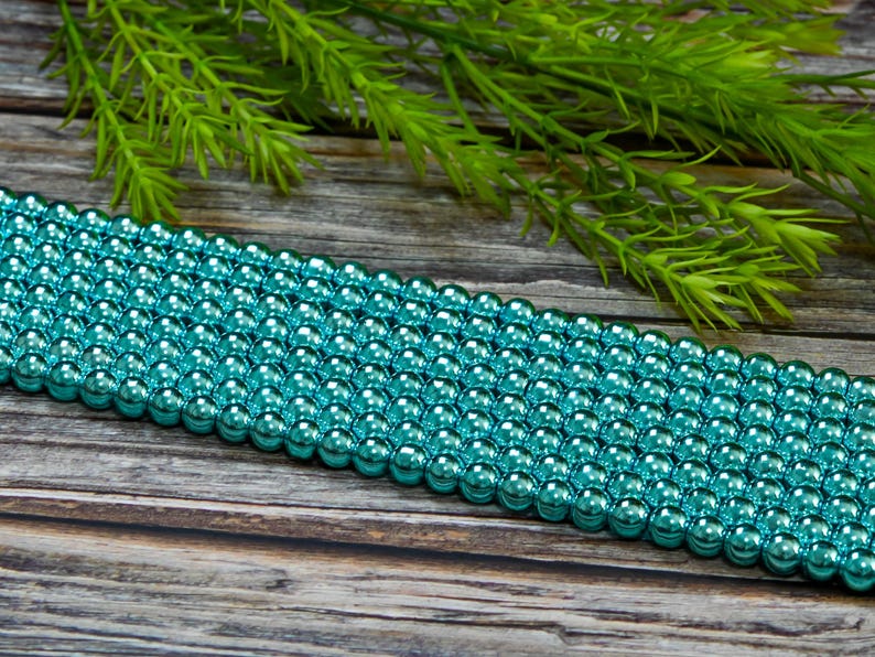 May include: A strand of turquoise metallic round beads. The beads are arranged in rows and have a shiny, reflective finish.