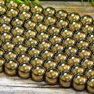 May include: A close-up of a strand of shiny gold beads. The beads are round and smooth, and they are arranged in a staggered pattern. The beads are on a wooden surface.