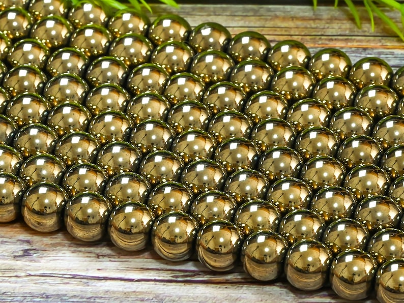 May include: A close-up of a strand of shiny gold beads. The beads are round and smooth, and they are arranged in a staggered pattern. The beads are on a wooden surface.