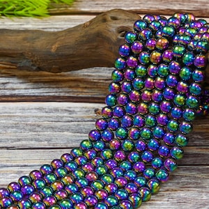 May include: A strand of iridescent hematite beads in a rainbow of colors, including purple, blue, green, and gold. The beads are round and smooth, and they are strung on a clear thread. The beads are displayed on a wooden surface.
