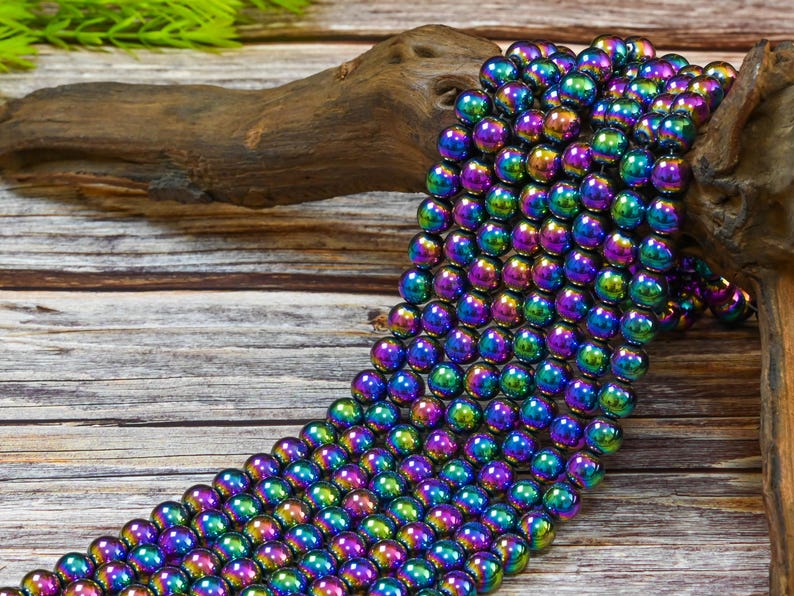 May include: A strand of iridescent hematite beads in a rainbow of colors, including purple, blue, green, and gold. The beads are round and smooth, and they are strung on a clear thread. The beads are displayed on a wooden surface.