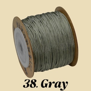 Puede incluir: Un carrete de cord&oacute;n gris. El cord&oacute;n est&aacute; enrollado alrededor de un carrete de cart&oacute;n con un n&uacute;cleo de pl&aacute;stico blanco. El texto "38. Gray" est&aacute; impreso en negro en la parte inferior de la imagen. El cord&oacute;n parece estar hecho de un material liso y brillante.