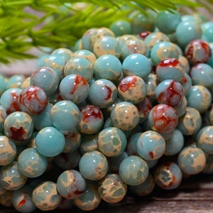 Può includere: Una collezione di perline rotonde e levigate con un motivo marmorizzato nei toni del blu turchese, crema e marrone rossastro intenso. Ogni perlina ha una finitura liscia e lucida con una rete distinta di venature e macchie sulla superficie.