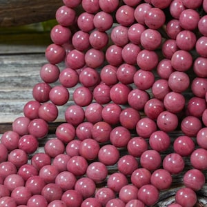 May include: Close-up of a collection of pink gemstone beads. The beads are round, with a polished surface and a marbled appearance. The beads are strung together, creating a cascading effect. The color is a deep, rosy pink.