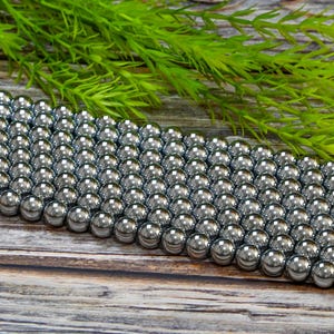 May include: A strand of silver-toned hematite beads. The beads are round and smooth, and they are arranged in a row on a wooden surface.