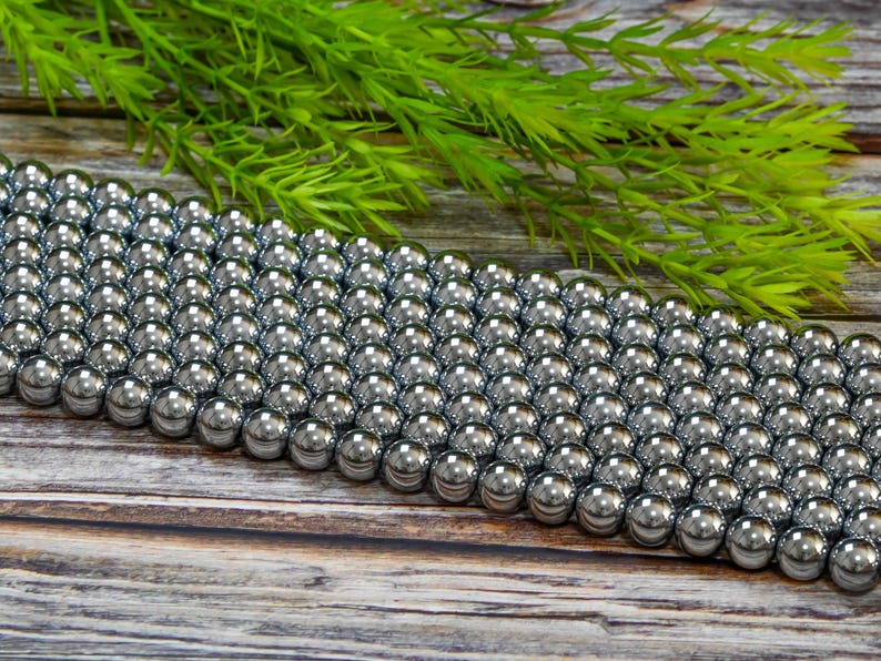 May include: A strand of silver-toned hematite beads. The beads are round and smooth, and they are arranged in a row on a wooden surface.