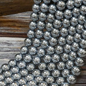 May include: A strand of silver-toned round beads, each bead is smooth and reflective. The beads are arranged in a cascading pattern, creating a visual texture.