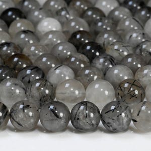 May include: A collection of clear, round beads with internal black inclusions. The beads vary in shades of gray and black, with a polished, reflective surface. The beads are arranged closely together, creating a textured pattern.