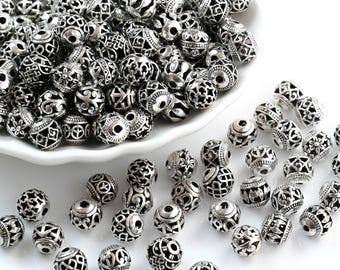 May include: Silver metal beads with detailed openwork patterns. The round beads display diverse designs, including floral, geometric, and abstract shapes. Some beads are in a white dish, others scattered.