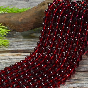 May include: A strand of smooth, round, deep red garnet beads arranged in multiple parallel rows. The translucent, dark crimson spheres are strung together on a thin pink cord, creating a uniform, polished appearance across the entire collection.