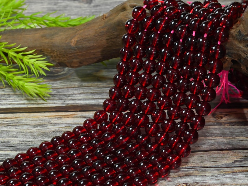 May include: A strand of smooth, round, deep red garnet beads arranged in multiple parallel rows. The translucent, dark crimson spheres are strung together on a thin pink cord, creating a uniform, polished appearance across the entire collection.