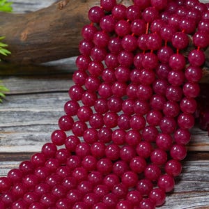 Peut inclure: Gros plan sur des rang&eacute;es de perles de pierres pr&eacute;cieuses rouge fonc&eacute;. Les perles rondes et polies sont enfil&eacute;es sur un cordon rouge. L'arri&egrave;re-plan pr&eacute;sente un morceau de bois et du feuillage vert, cr&eacute;ant une esth&eacute;tique naturelle. Chaque perle mesure environ 0,8 cm de diam&egrave;tre.
