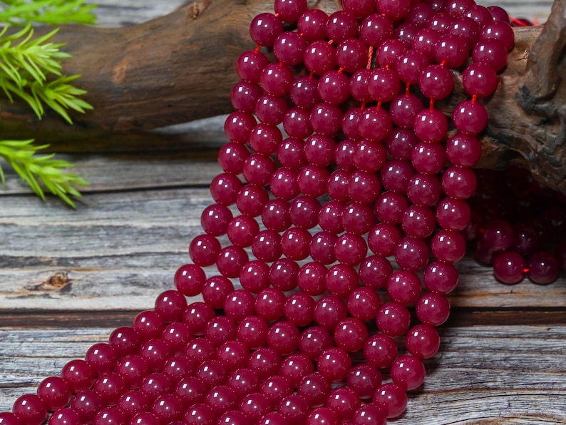 Peut inclure: Gros plan sur des rang&eacute;es de perles de pierres pr&eacute;cieuses rouge fonc&eacute;. Les perles rondes et polies sont enfil&eacute;es sur un cordon rouge. L'arri&egrave;re-plan pr&eacute;sente un morceau de bois et du feuillage vert, cr&eacute;ant une esth&eacute;tique naturelle. Chaque perle mesure environ 0,8 cm de diam&egrave;tre.