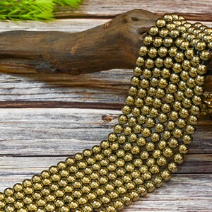May include: A strand of gold hematite beads, each bead is round and smooth. The beads are arranged in a loose pile on a wooden surface.