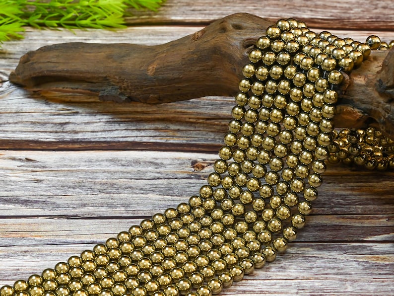 May include: A strand of gold hematite beads, each bead is round and smooth. The beads are arranged in a loose pile on a wooden surface.