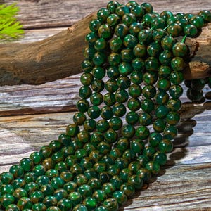 Puede incluir: Una hebra de cuentas de piedras preciosas verdes y doradas. Las cuentas son redondas y tienen un patr&oacute;n moteado. Las cuentas est&aacute;n enhebradas en un cord&oacute;n y est&aacute;n sobre una superficie de madera.