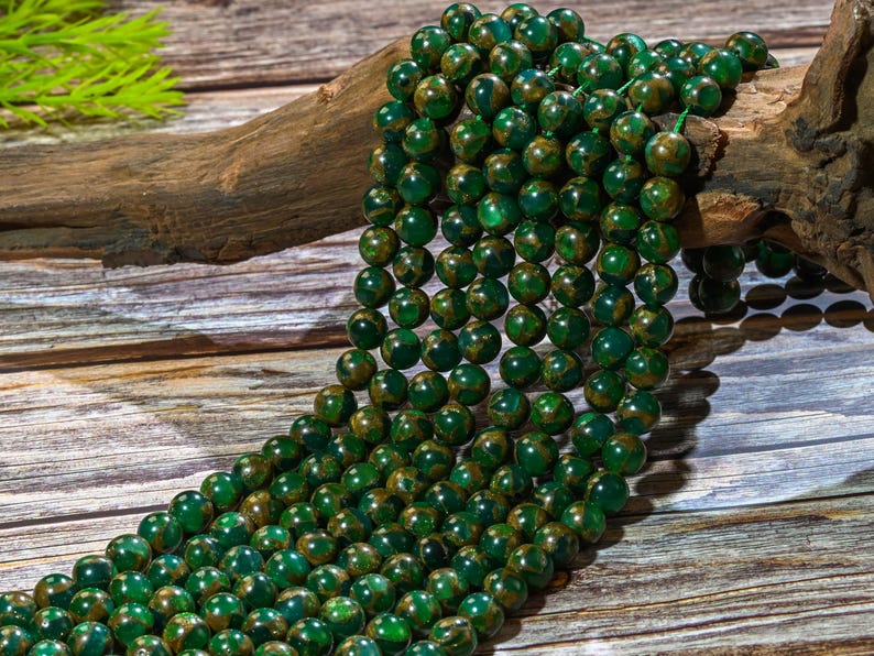 Puede incluir: Una hebra de cuentas de piedras preciosas verdes y doradas. Las cuentas son redondas y tienen un patr&oacute;n moteado. Las cuentas est&aacute;n enhebradas en un cord&oacute;n y est&aacute;n sobre una superficie de madera.