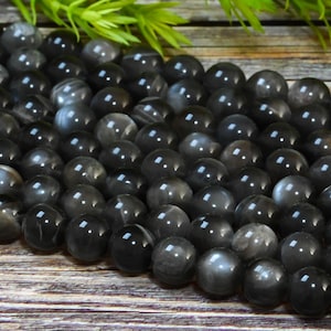 Pode incluir: Um fio de contas de pedras preciosas pretas com um efeito brilhante e iridescente. As contas são redondas e lisas, e estão dispostas em uma visão de close-up.