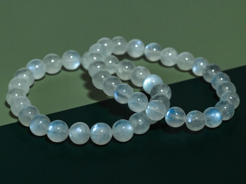 Puede incluir: Dos pulseras el&aacute;sticas compuestas por cuentas redondas y transl&uacute;cidas de piedra lunar. Cada cuenta presenta una superficie lisa y pulida con una sutil adularescencia azul iridiscente que capta la luz, creando un brillo suave en las piedras.