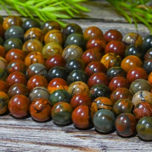 May include: A collection of polished, round gemstones in shades of red, brown, green, and yellow. The beads are arranged closely together, showcasing their varied patterns and colors. The gemstones are likely used for jewelry making or crafting.