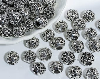 May include: A collection of silver filigree beads with intricate openwork patterns, such as floral and geometric designs. The spherical beads are displayed on a white surface and in a white dish. These decorative beads are suitable for jewellery making.