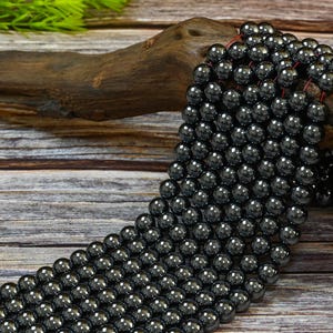 May include: A strand of black hematite beads, each bead is round and smooth. The beads are strung on a thin cord and are displayed on a piece of driftwood.